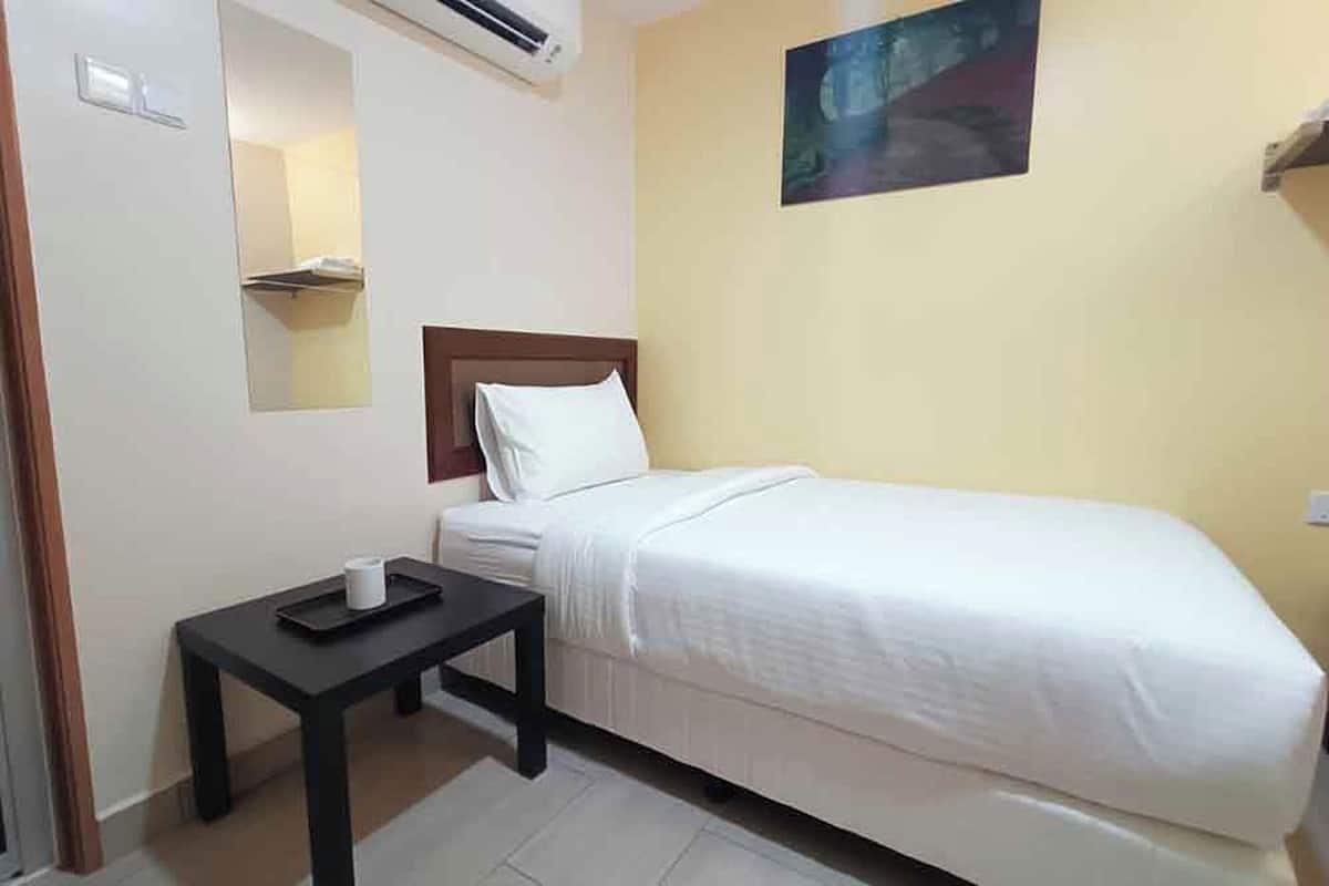 A single bed with a white comforter, a small bedside table with a tray, a wall-mounted air conditioner, a painting, and a light switch.