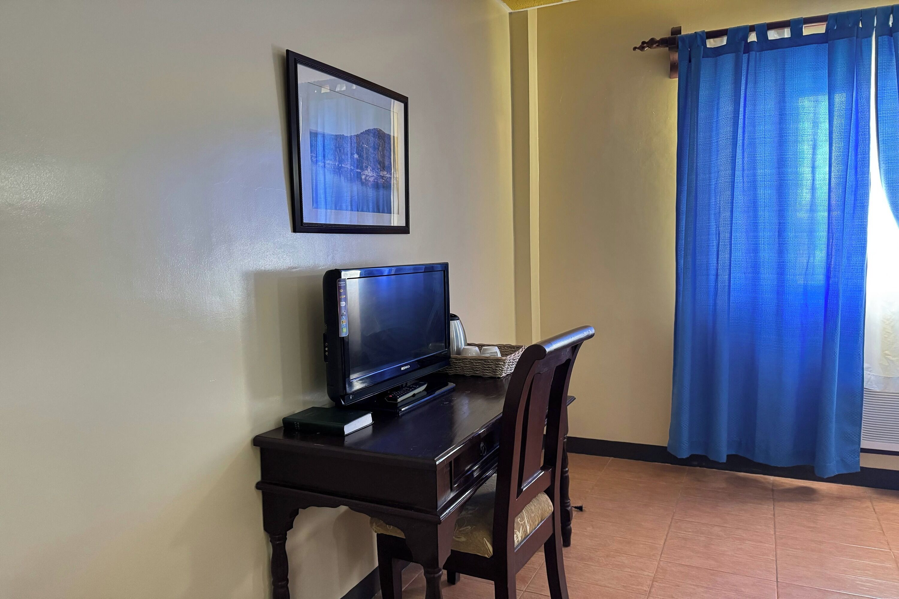 Una habitación con un escritorio de madera oscura, televisor, un cuadro en la pared y una cortina azul.