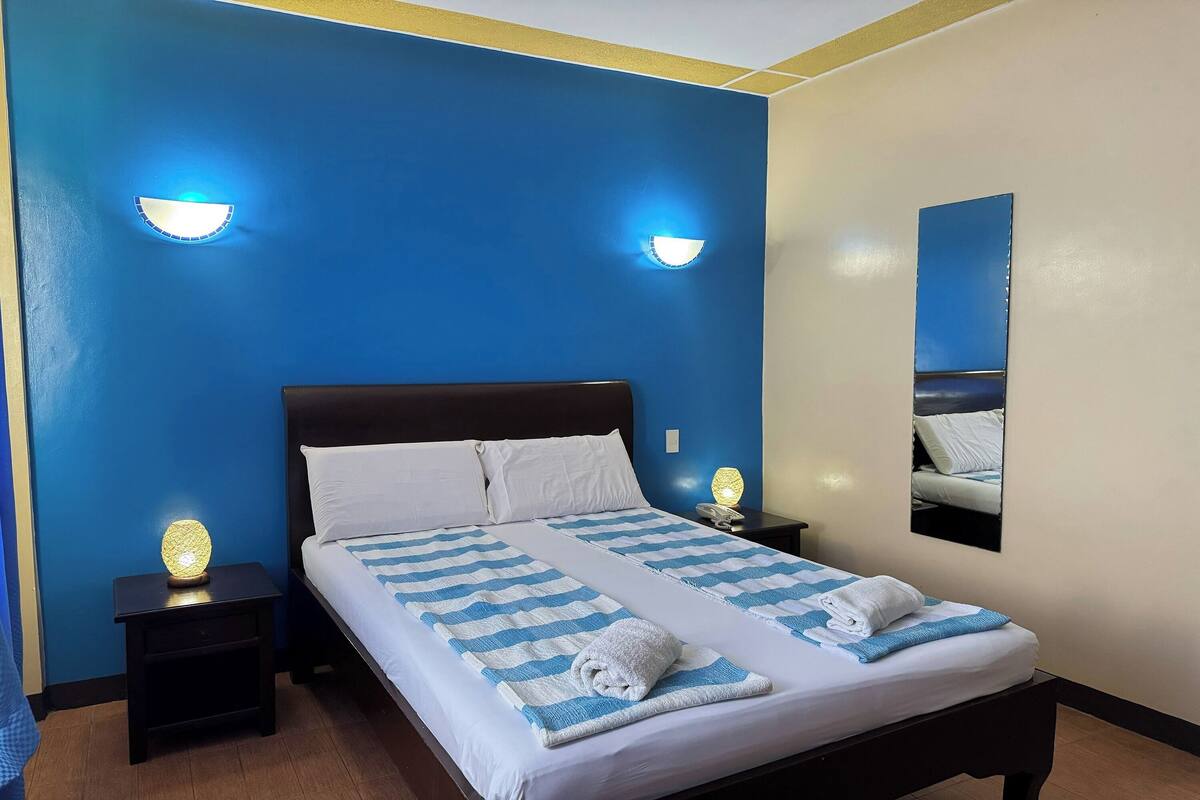 Ein ordentlich bezogenes Bett mit einer gestreiften Decke und aufgerollten Handtüchern, zwei Nachttische mit Lampen und ein Spiegel an der Wand.