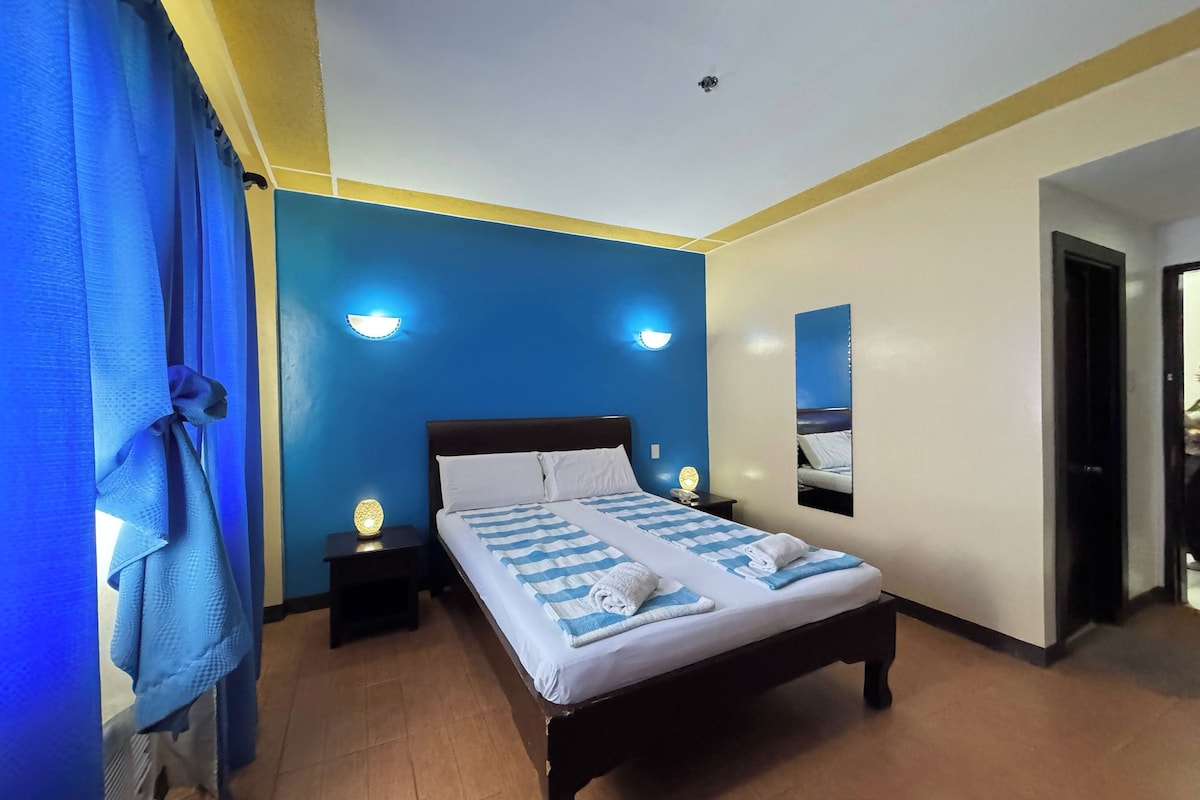 Una cama doble con una colcha azul y blanca a rayas, dos lámparas de noche y un espejo fijado a la pared.