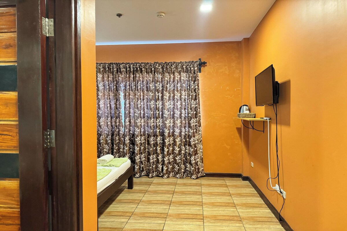 Une chambre avec un lit, des rideaux, un téléviseur fixé au mur et une étagère avec une bouilloire.