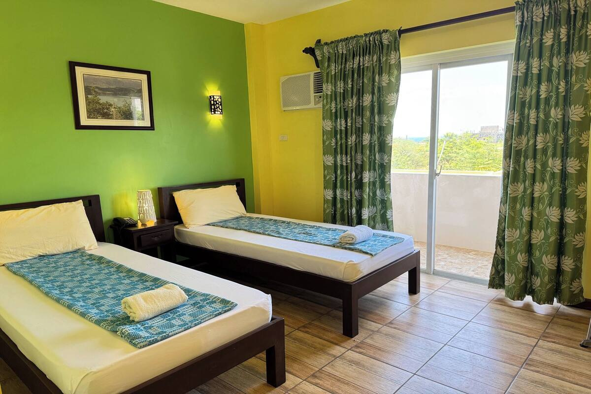 Ein Hotelzimmer mit zwei Betten, einer grünen Wand, einem gerahmten Bild, einem Nachttisch und einem Fenster mit Vorhängen.