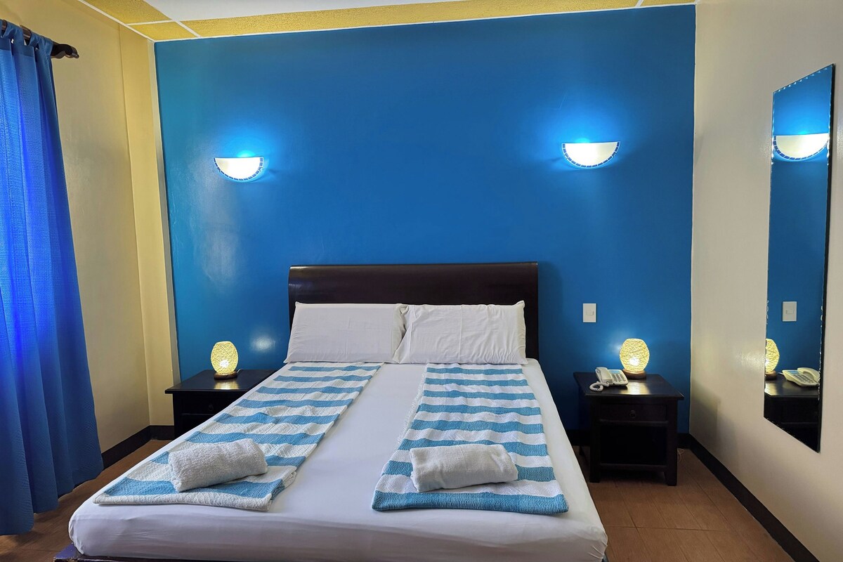 Une chambre d’hôtel avec un lit, deux tables de chevet, une commode, un miroir et un rideau.