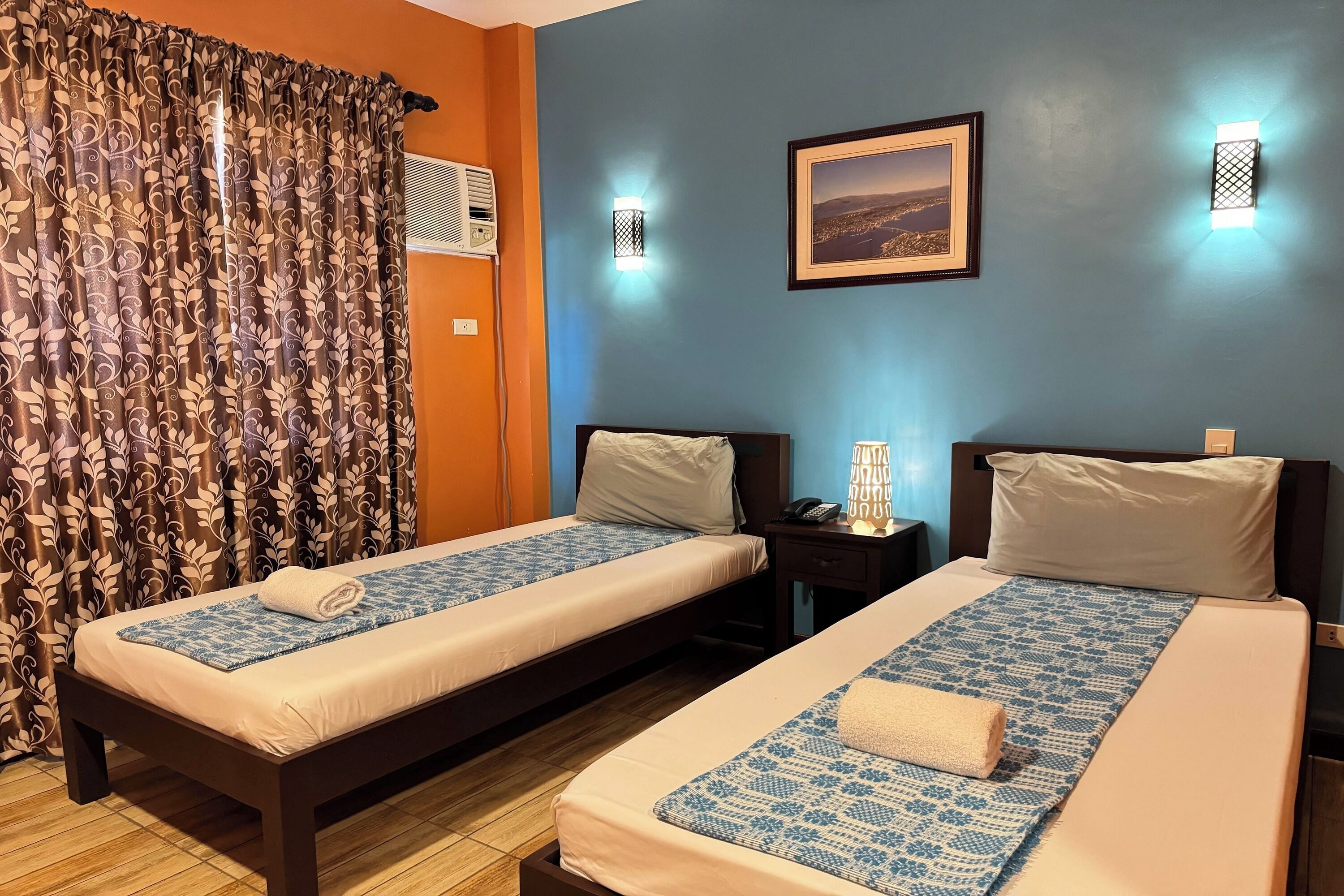 Una habitación de hotel con dos camas, una ventana con cortinas, un aire acondicionado instalado en la pared y dos apliques.