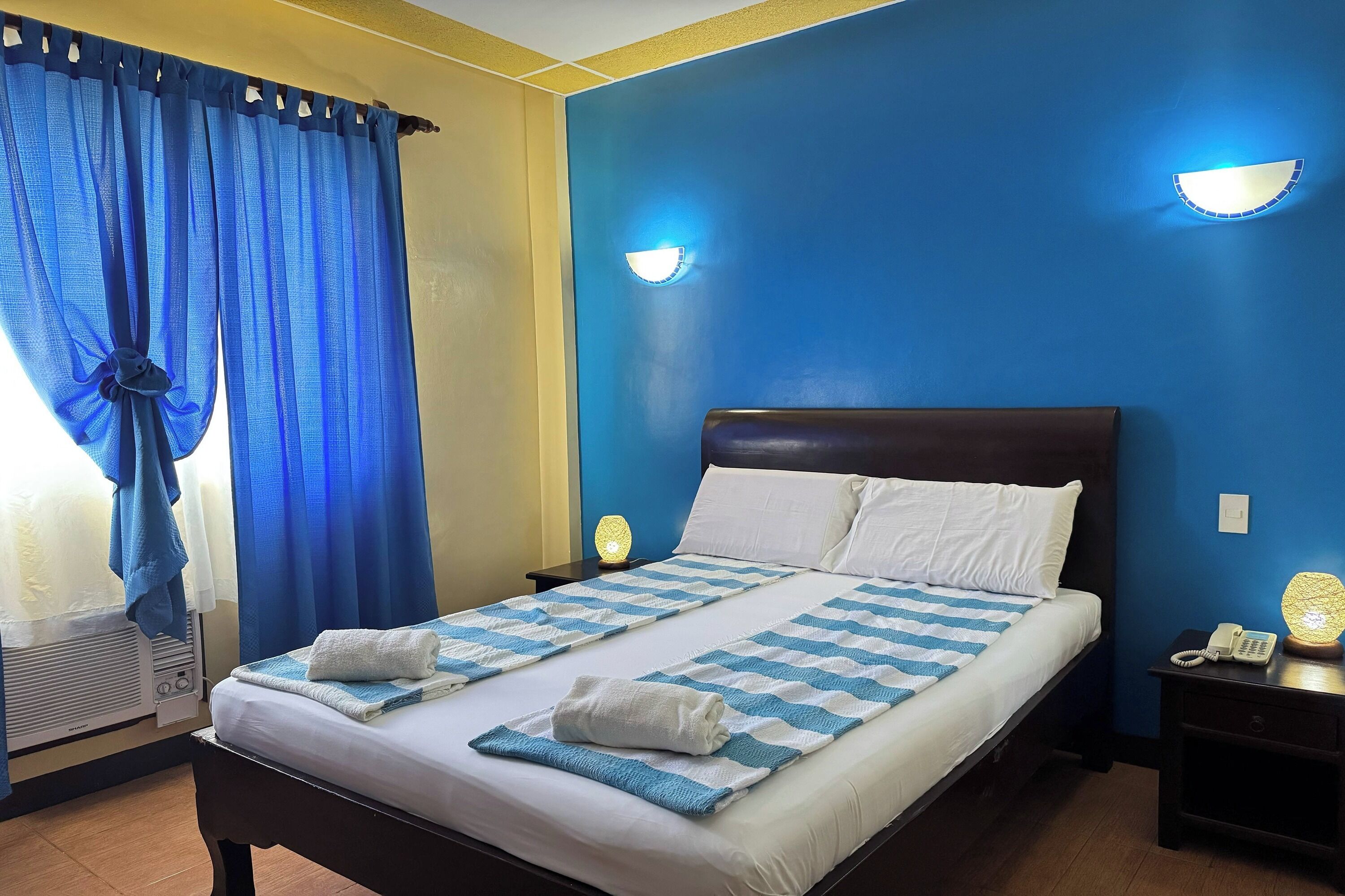 A double bed with white linens, a blue and white striped blanket, and two towels. There are two bedside tables with lamps, a wall-mounted light fixture, and a telephone on the nightstand.