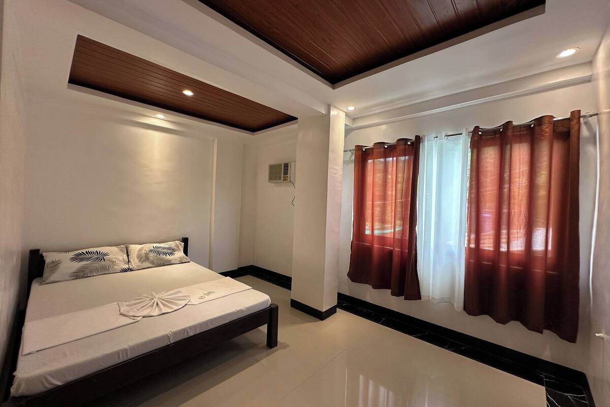Une chambre à coucher avec un lit, un plafond en bois, des murs blancs et une fenêtre avec des rideaux.