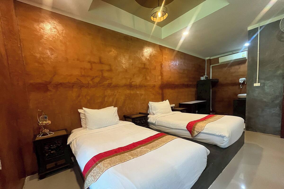 A hotel room with two beds, a small table, a nightstand, a wall-mounted air conditioner, and a ceiling light fixture.