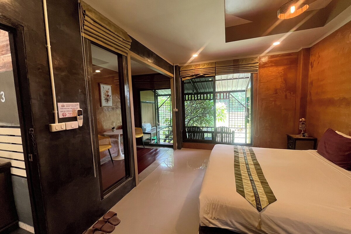 Une chambre d’hôtel avec un grand lit, une petite table avec des chaises, une porte avec un téléphone et une pancarte, et une vue sur l’extérieur par une fenêtre.