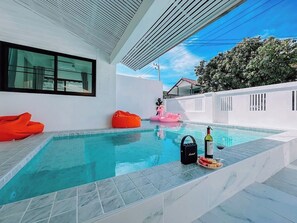 A modern outdoor pool area with a clear blue pool, a lounge chair, a pink inflatable float, and a bottle of wine on the poolside. - Baan Talay Home Jomtien Beach (Pattaya)