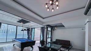 Ein modernes Zimmer mit Billardtisch, einem Glastisch mit Stühlen, einer Couch und einem großen Fenster mit Blick auf die Stadt.
