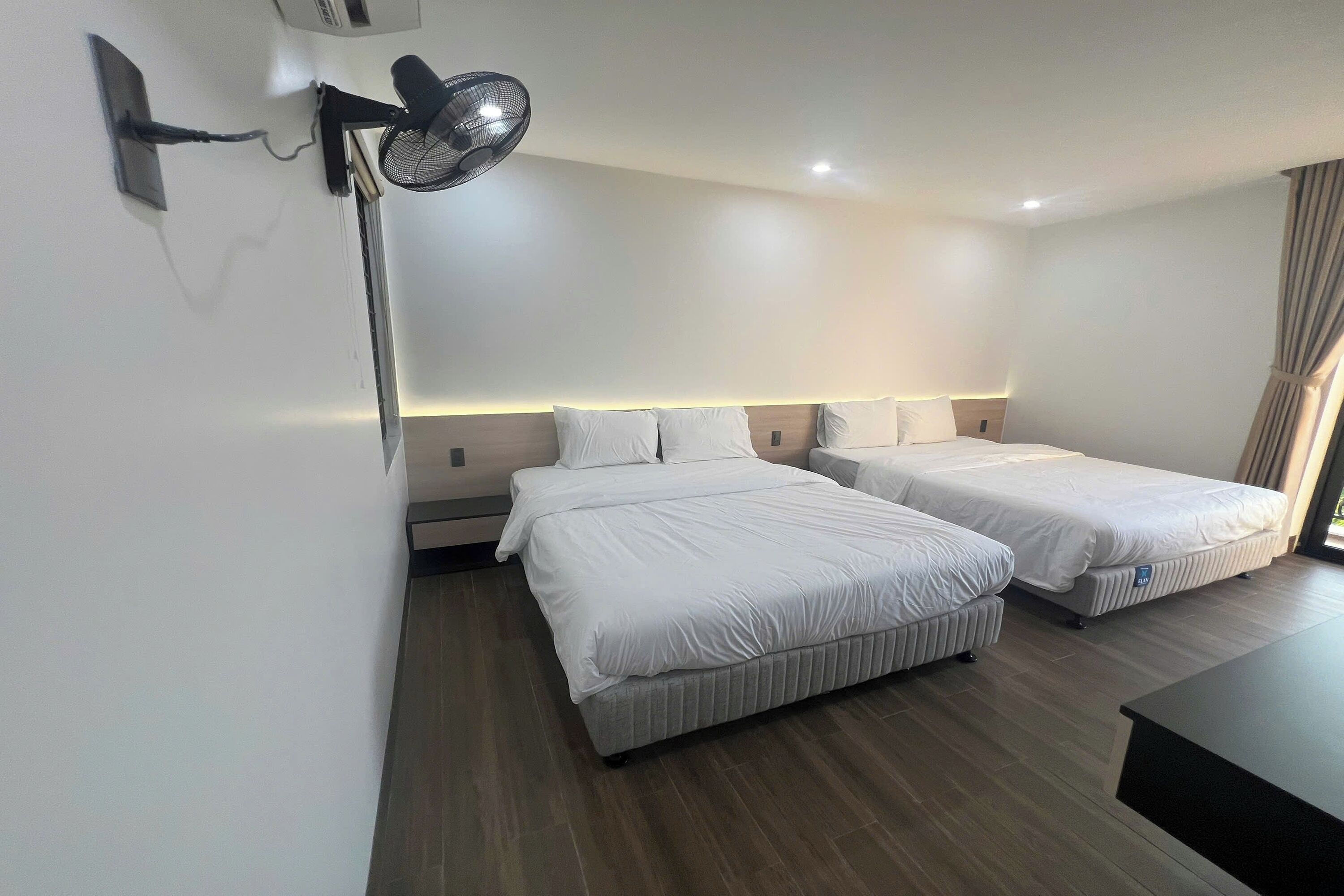 Hotellihuone, jossa on kaksi sänkyä, työpöytä, seinään kiinnitetty tuuletin ja kattovalaisin.