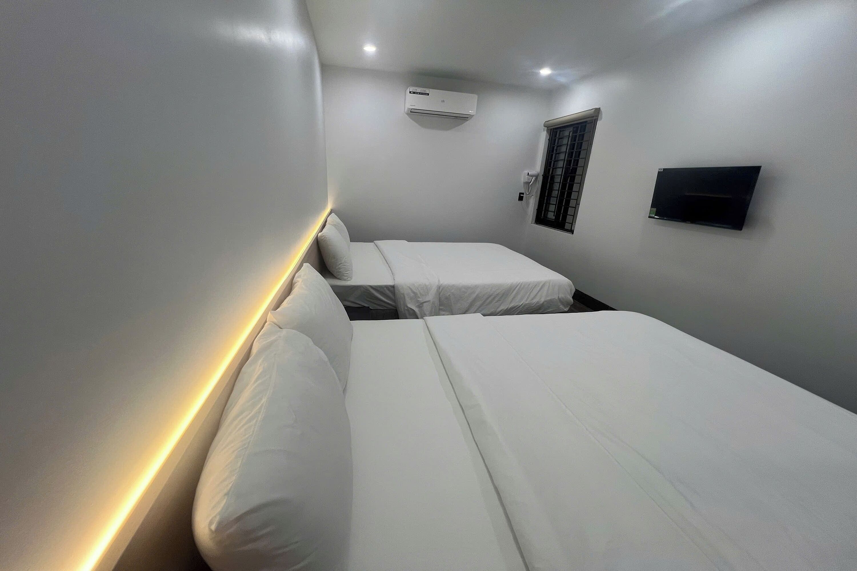 A hotel room with two single beds, a wall-mounted air conditioner, a window with blinds, and a flat-screen TV.
