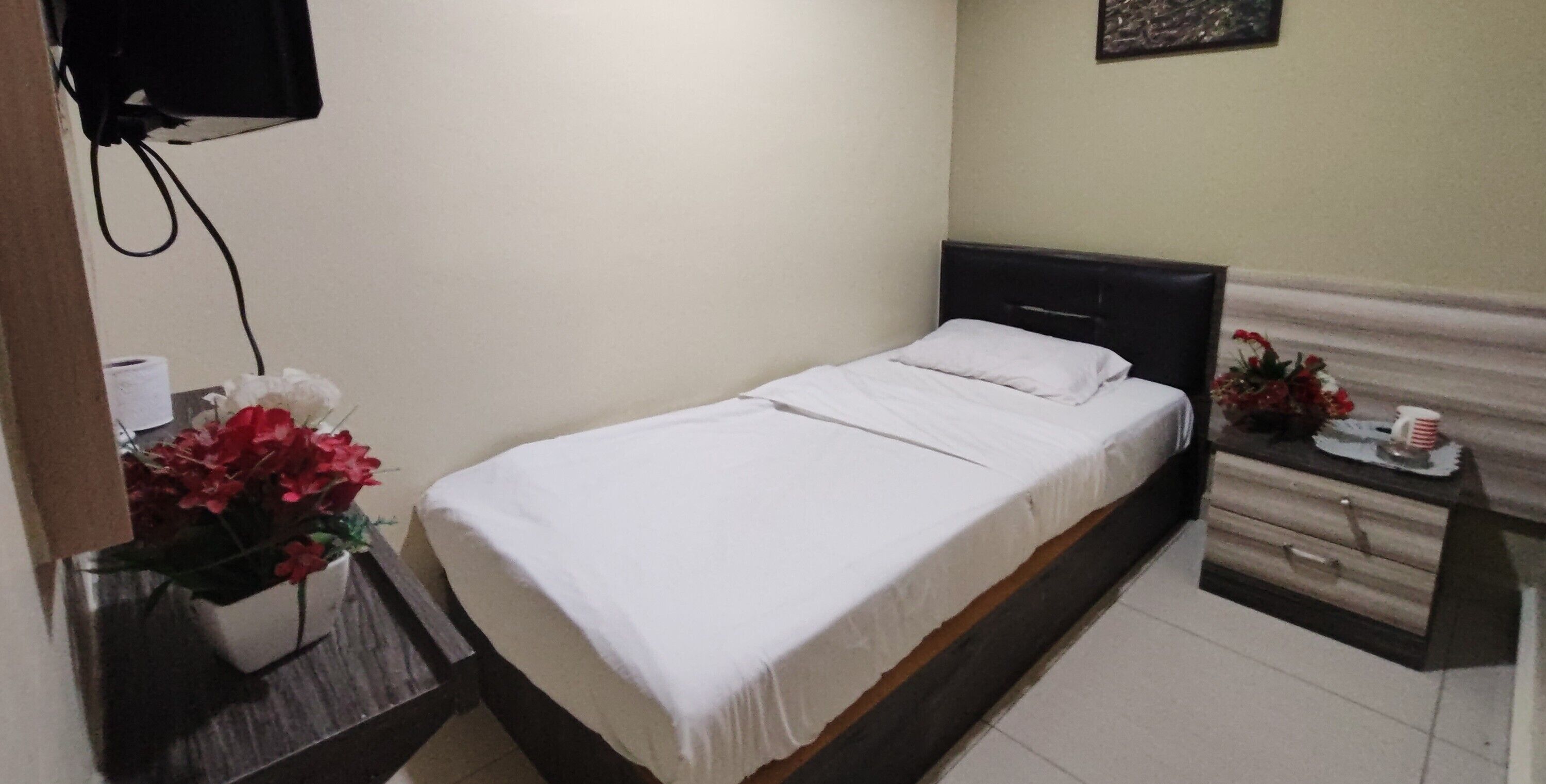 A single bed with a white bedspread and a wooden headboard. There is a nightstand with a vase of red flowers and a small table with a cup and a plate.