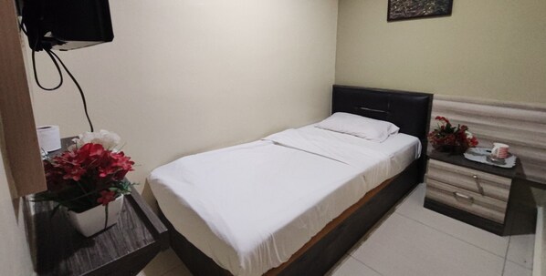 A single bed with a white bedspread and a wooden headboard. There is a nightstand with a vase of red flowers and a small table with a cup and a plate.