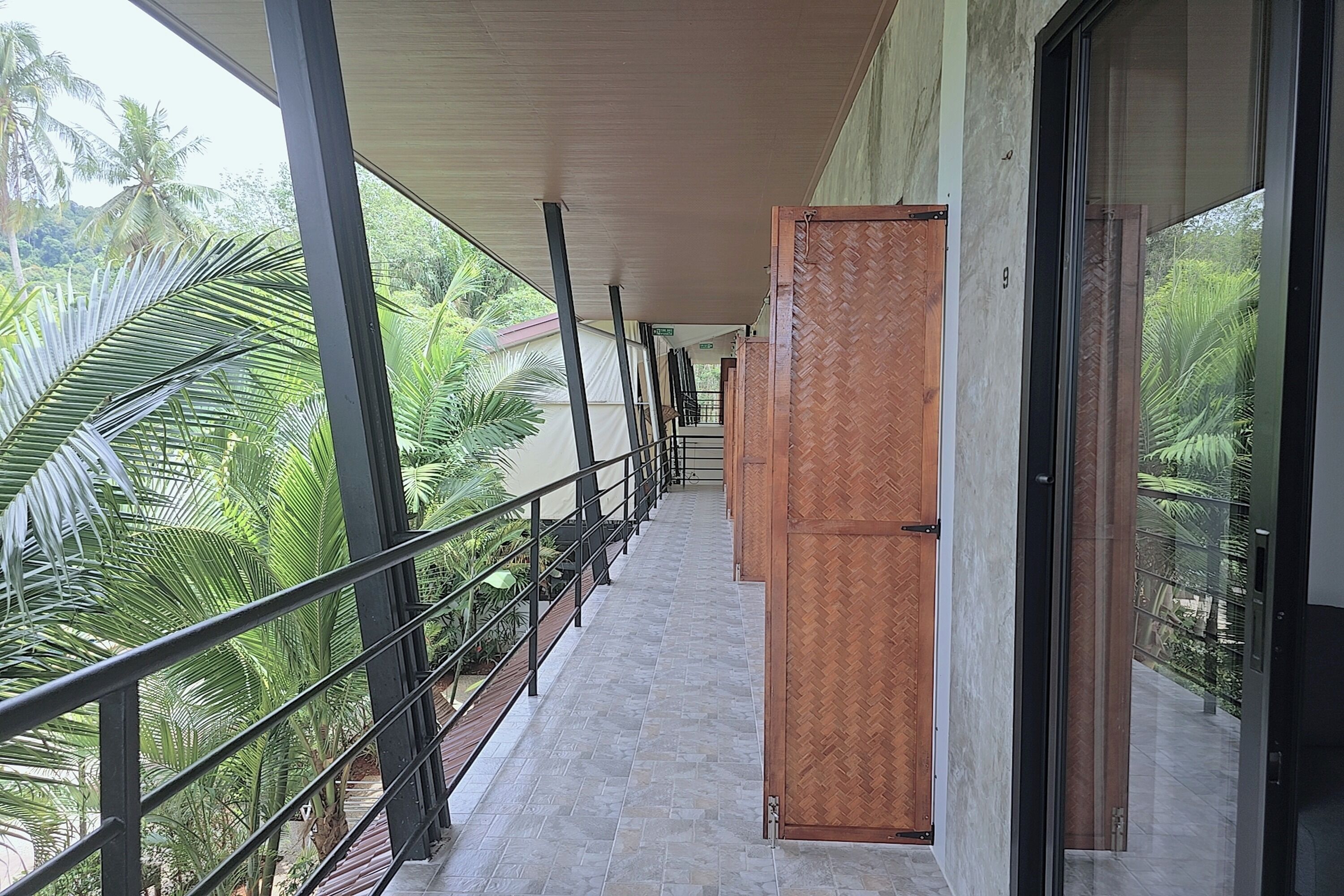 A covered corridor with a metal railing, a wooden door, and a view of palm trees.