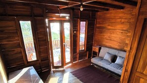 A cozy cabin interior with a wooden floor, a sofa, a small table, and a window with a view of the outdoors. - Munduk Terrace (Munduk)