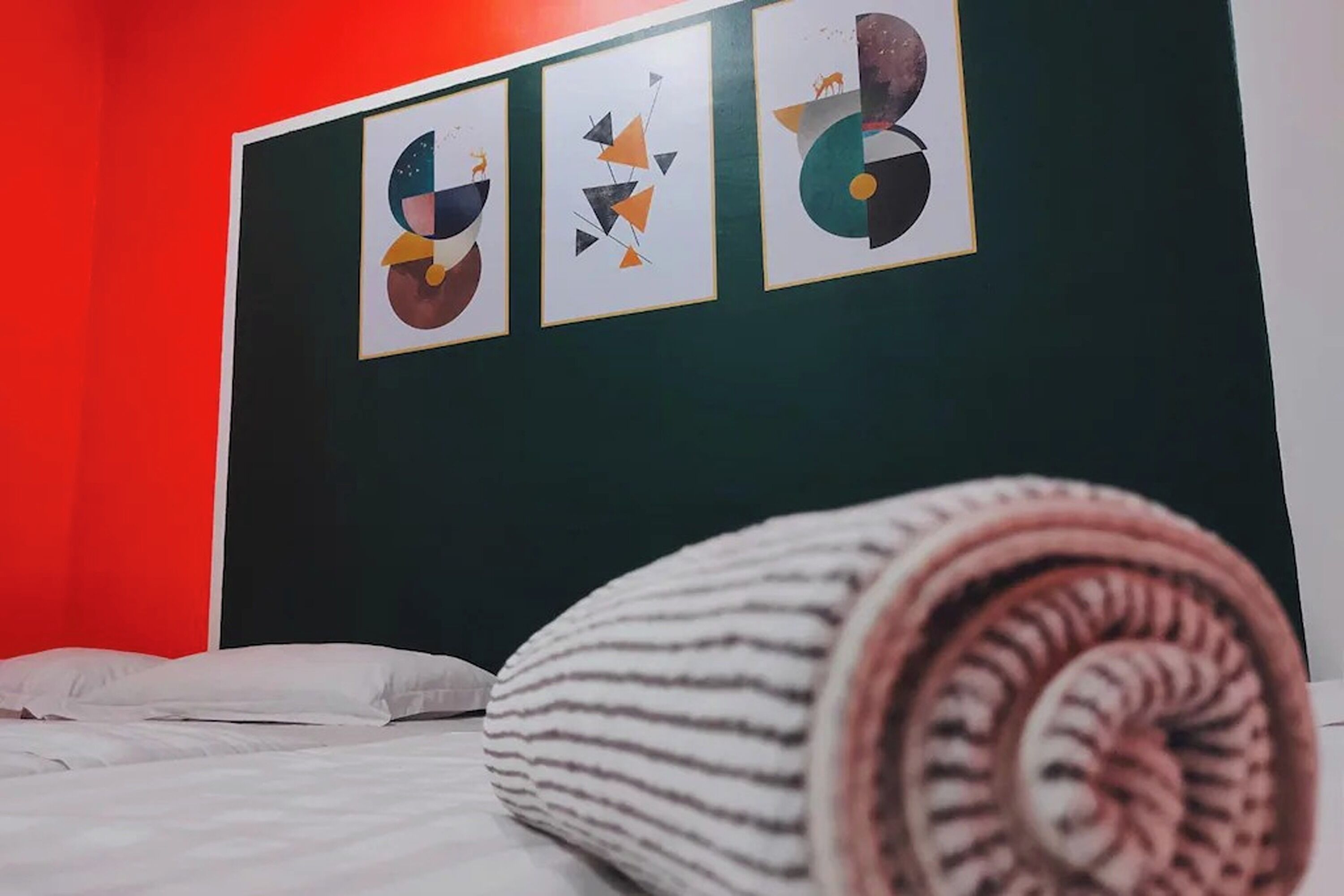 A bed with white bedding and a rolled-up striped blanket in a room with a red accent wall and three abstract artwork pieces on the wall.