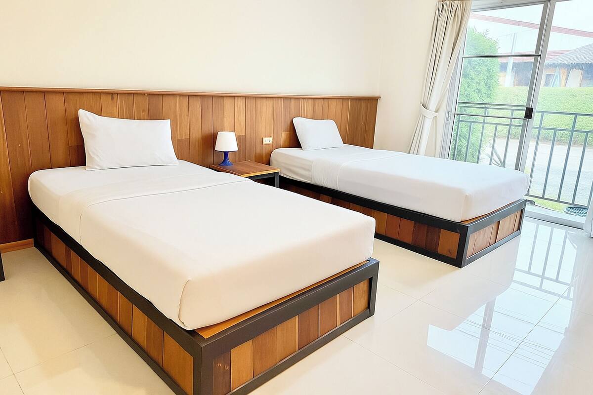 A hotel room with two beds, wooden headboard, white bedding, a nightstand with a lamp, and a balcony with a view.