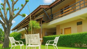 Un bâtiment à deux étages doté d’un balcon, avec un arbre et deux chaises blanches dans un espace aménagé en jardin.