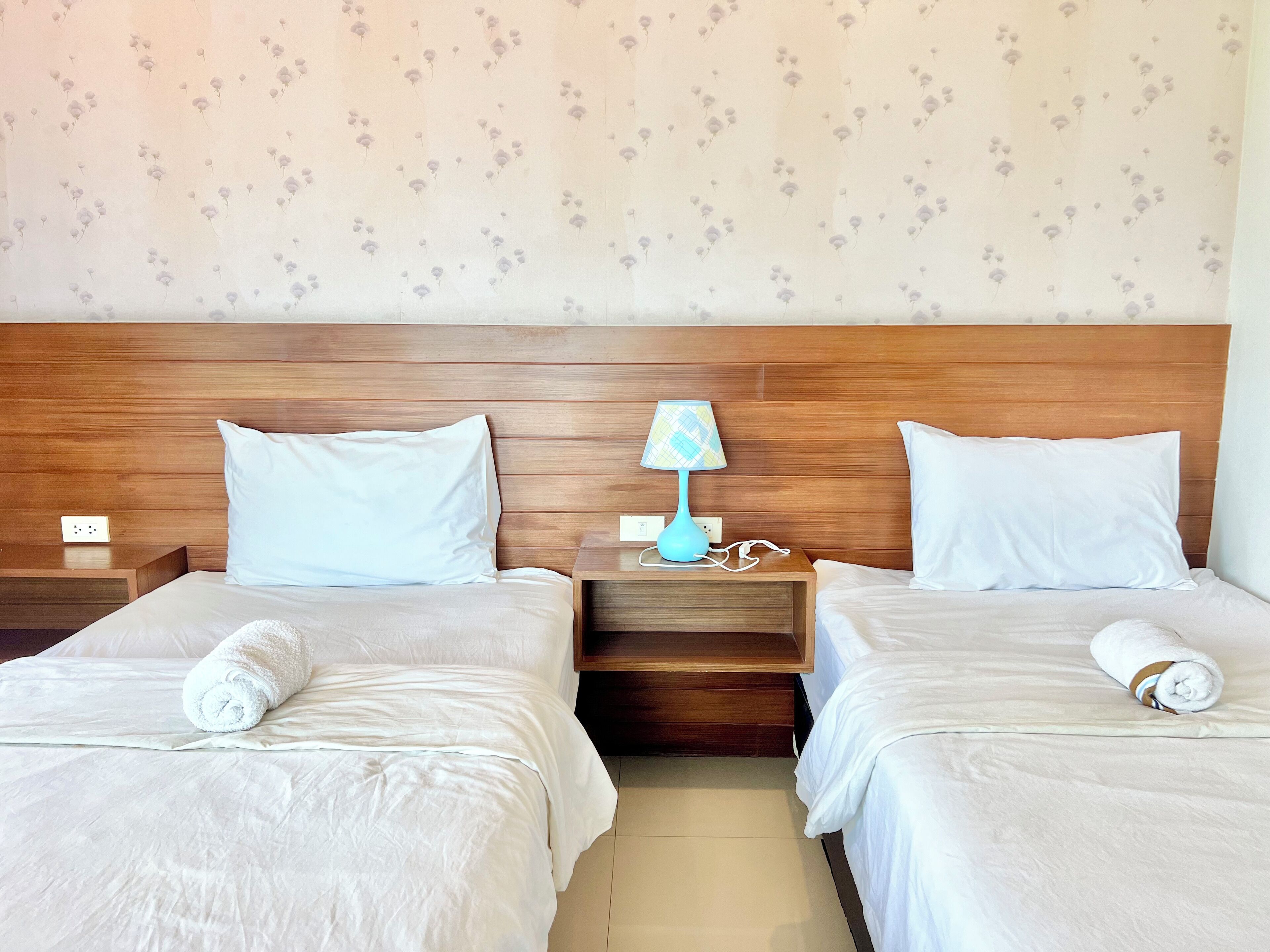 A hotel room with two beds, wooden headboards, white bedding, a nightstand with a lamp, and a floral wallpaper pattern.