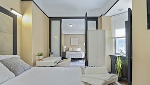 A hotel room with a large bed, a desk, a chair, a small table with a plant, and a window with blinds. - Chic Two Bedroom Apartment at the Historic Mansfield Residence (New York)