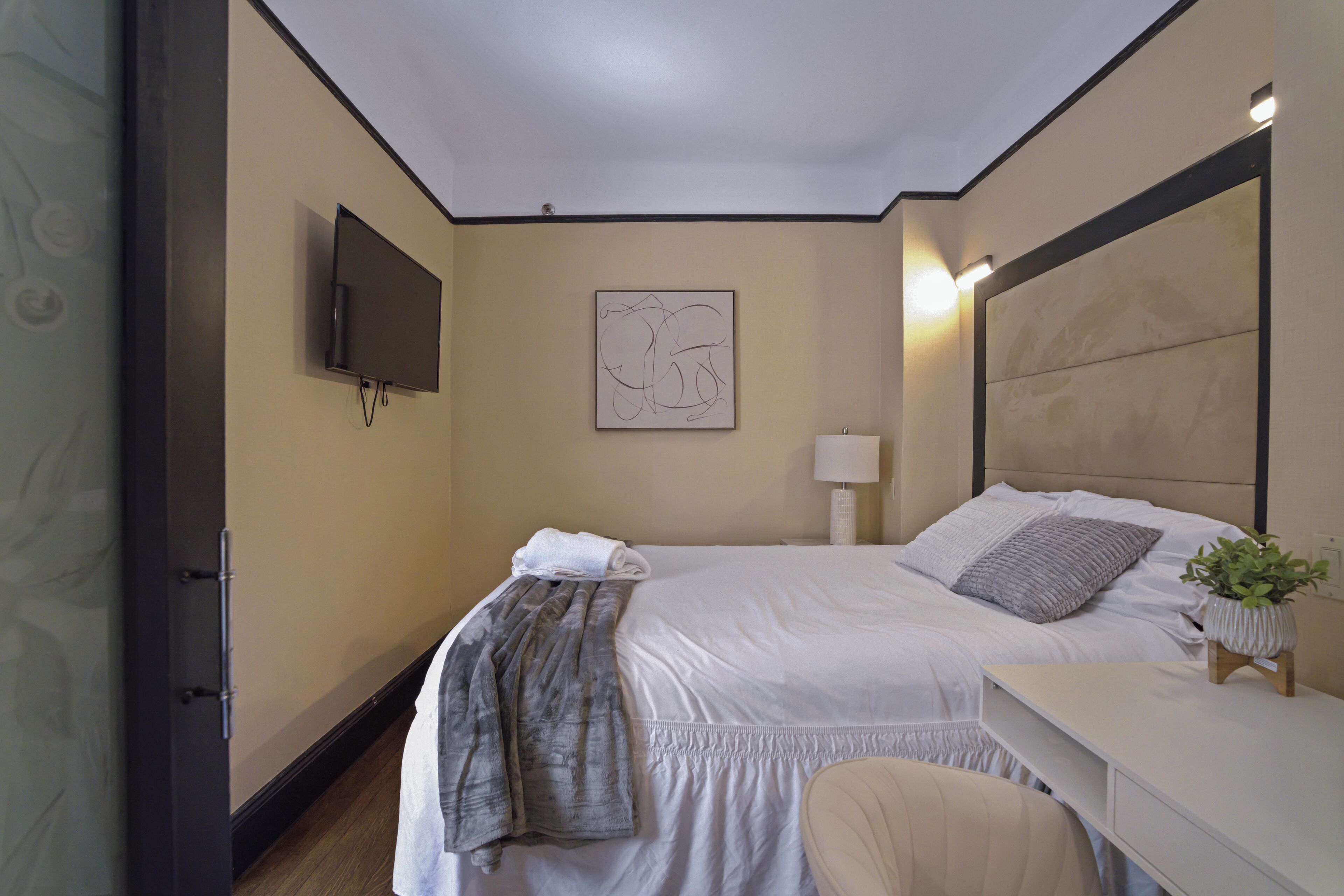 Une chambre d’hôtel moderne avec un grand lit, un téléviseur à écran plat fixé au mur, une table de chevet avec une lampe et une petite plante en pot.