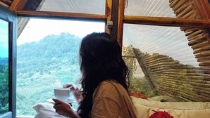 Una persona sentada en una cama dentro de una habitación con un ventanal grande, observando un paisaje montañoso.