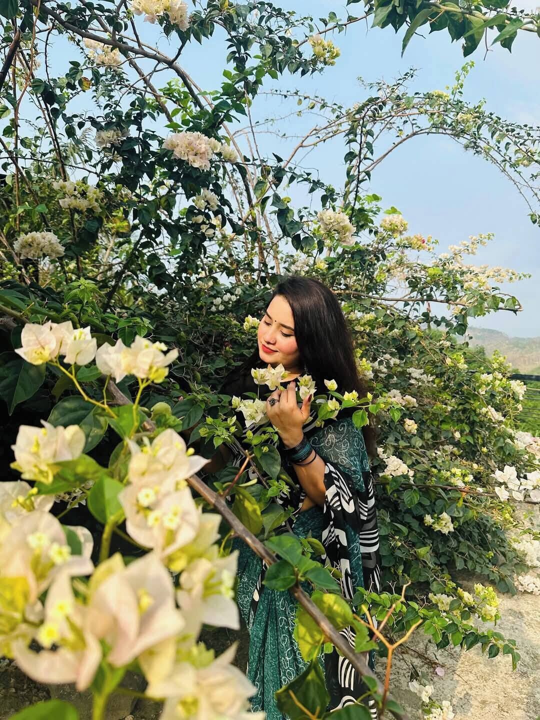 A woman standing among blooming white flowers, surrounded by lush greenery.
