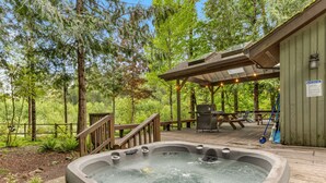 A hot tub on a wooden deck surrounded by trees, with a covered seating area and a picnic table. - Cozy Riverfront Retreat w/ Hot Tub (Rhododendron)