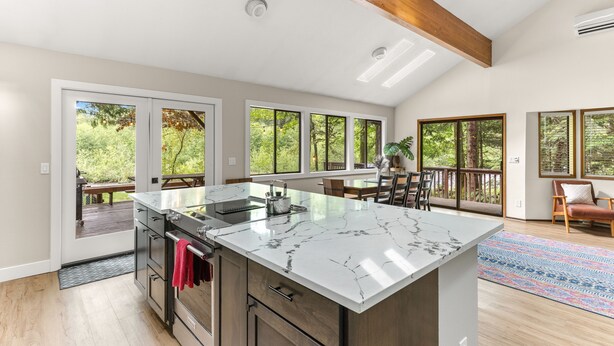 A spacious kitchen with a large island, wooden beams, and large windows offering views of the outdoors. - Cozy Riverfront Retreat w/ Hot Tub (Rhododendron)