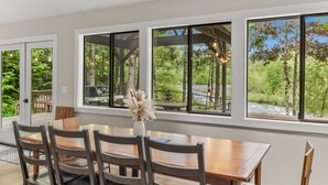 A dining area with a wooden table and chairs, large windows with a view of greenery and a wooden deck outside. - Cozy Riverfront Retreat w/ Hot Tub (Rhododendron)