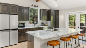 A modern kitchen with dark wooden cabinets, a white marble island with bar stools, and a view of the outdoors through large windows. - Cozy Riverfront Retreat w/ Hot Tub (Rhododendron)