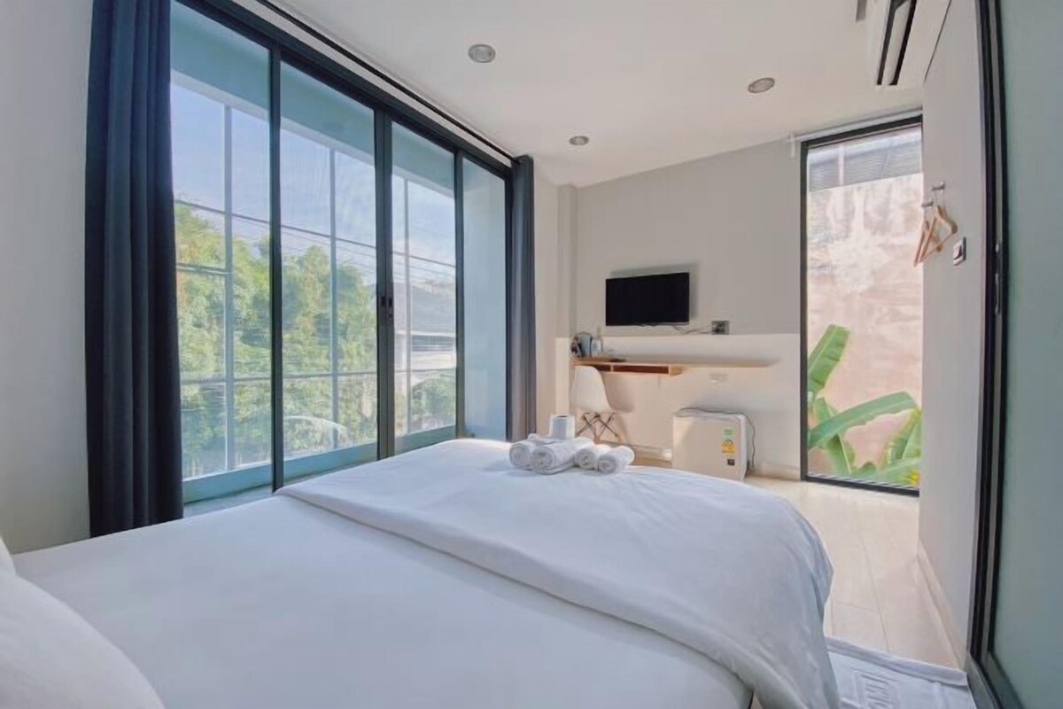 A modern hotel room with a large bed, a flat-screen TV mounted on the wall, and a balcony with a view of greenery.