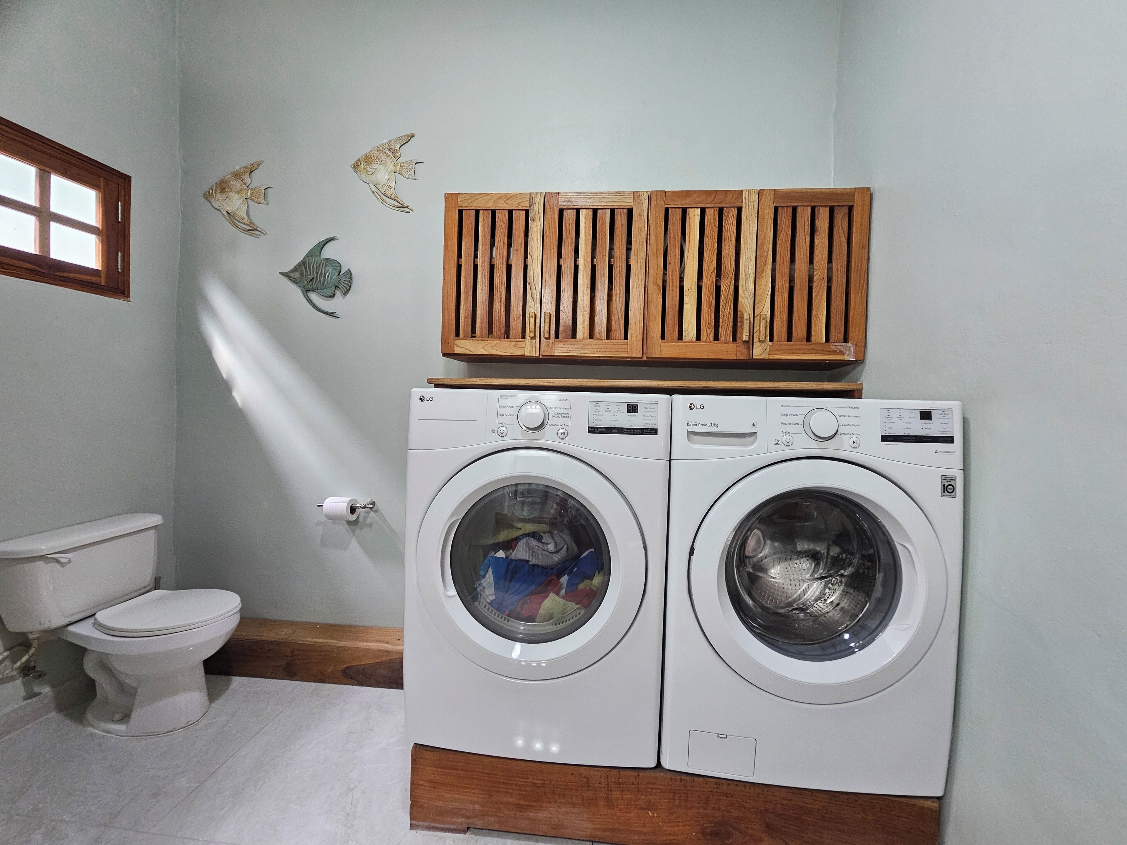 Une buanderie équipée de deux machines à laver LG à entraînement direct de 20 kg, de placards en bois au-dessus, de toilettes et de poissons décoratifs accrochés au mur.