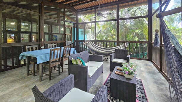 A covered outdoor seating area with wicker furniture, a hammock, and large windows offering a view of greenery. - Pedasi Craftsman Home, Large Screened Deck, Walk to Town (Los Santos Province)