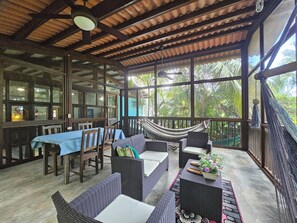 Un espace extérieur aménagé, abrité et doté de mobilier en osier, d’un hamac et de grandes fenêtres offrant une vue sur la verdure.