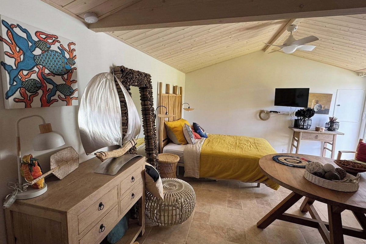 A cozy bedroom with a bed, bedside table, and a large mirror. There is a wooden dresser with drawers and a lamp. A dining table with chairs is visible in the foreground.