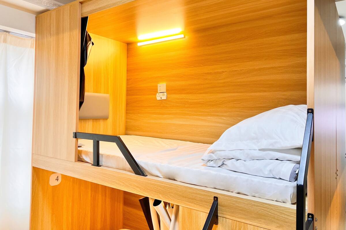 A bunk bed with a wooden finish, a bedside table, a lamp, and a wall socket.
