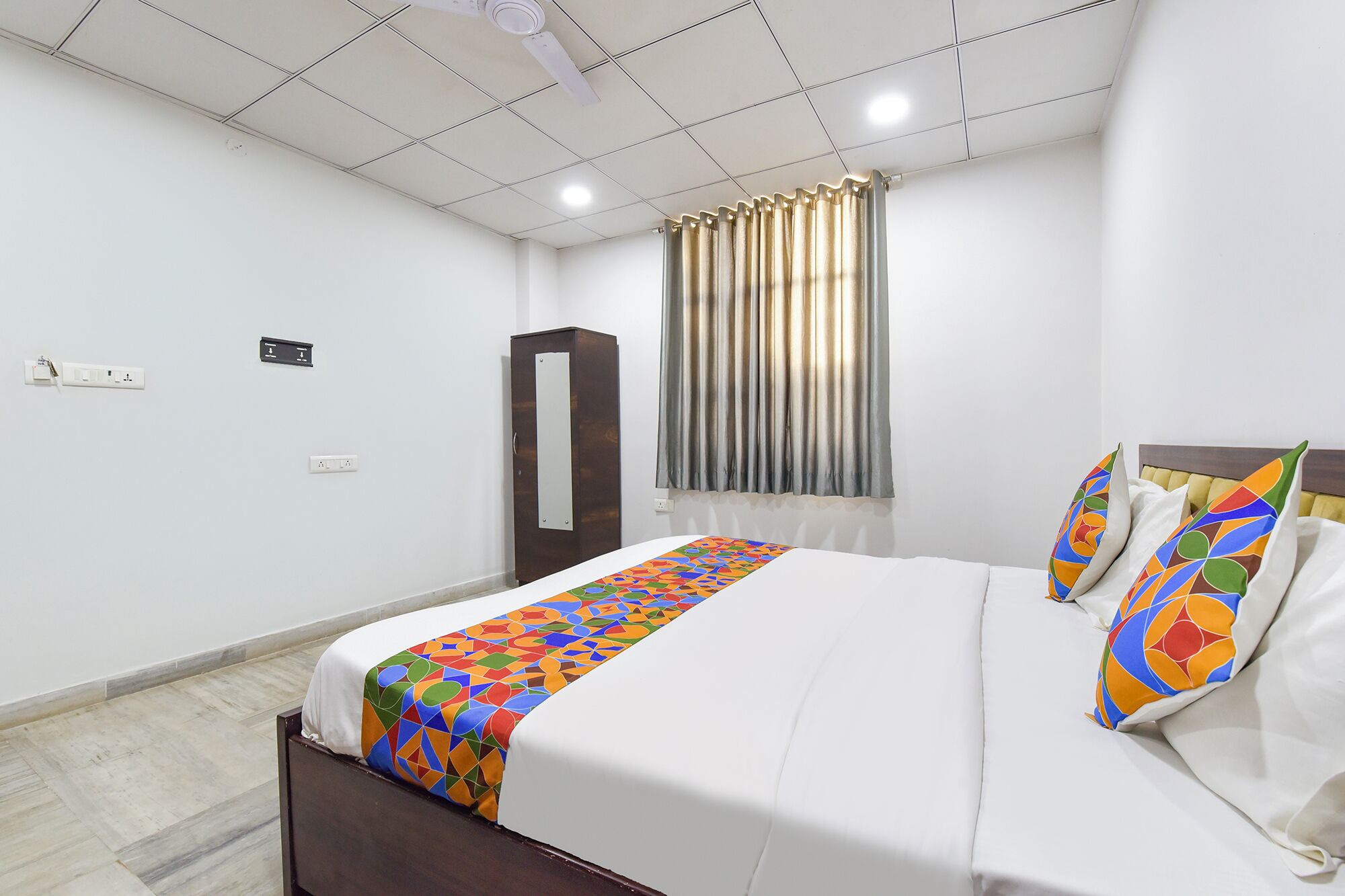 A hotel room with a bed, a wardrobe, a window with curtains, and a ceiling fan.