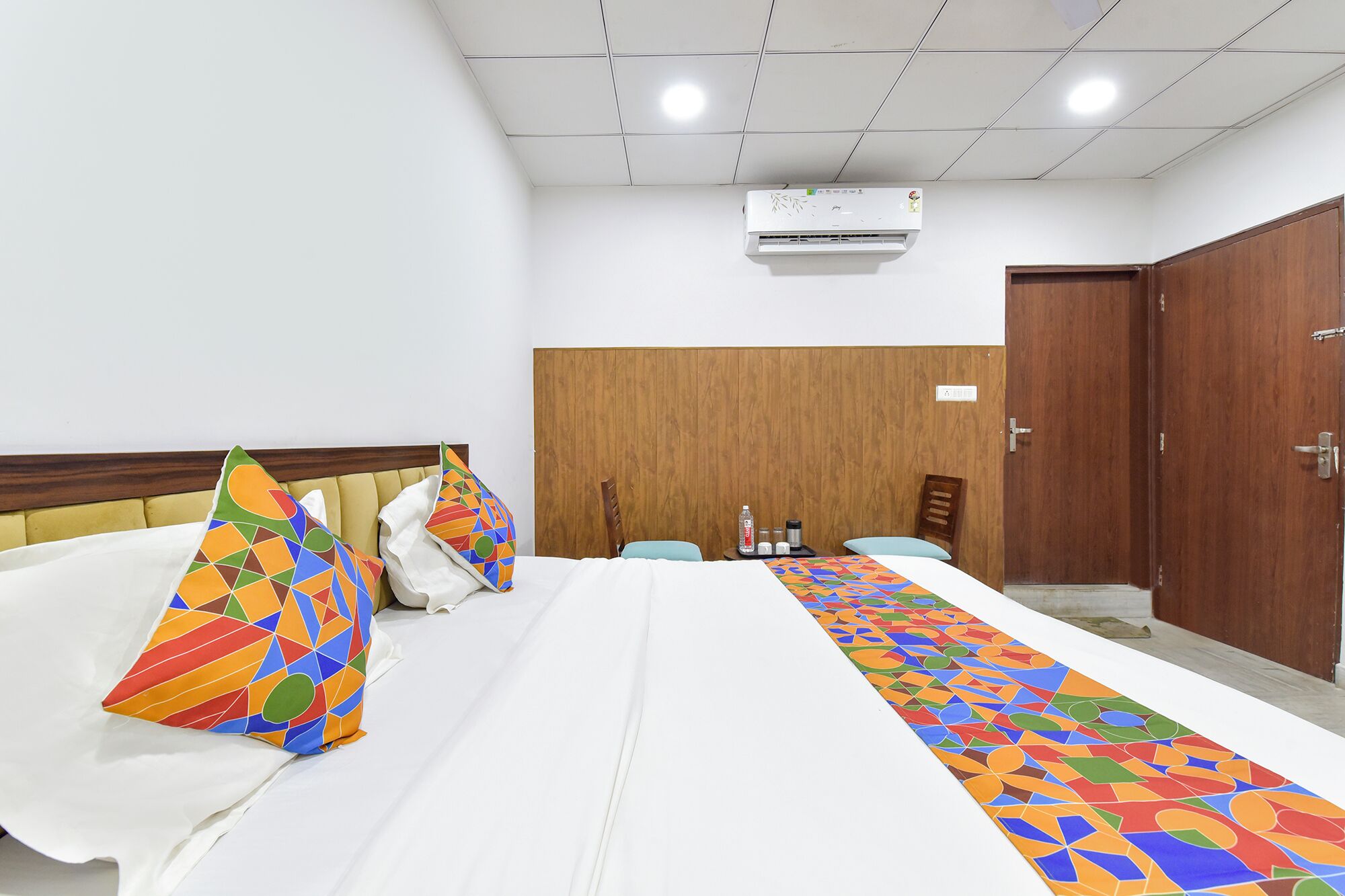 A hotel room with a bed, a wooden wardrobe, a desk with a chair, and an air conditioning unit on the ceiling.
