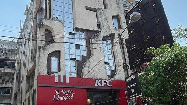Ein KFC Restaurant mit rotem und weißem Schild, in einem Gebäude mit vielen Fenstern, rechts daneben eine große schwarze Struktur.