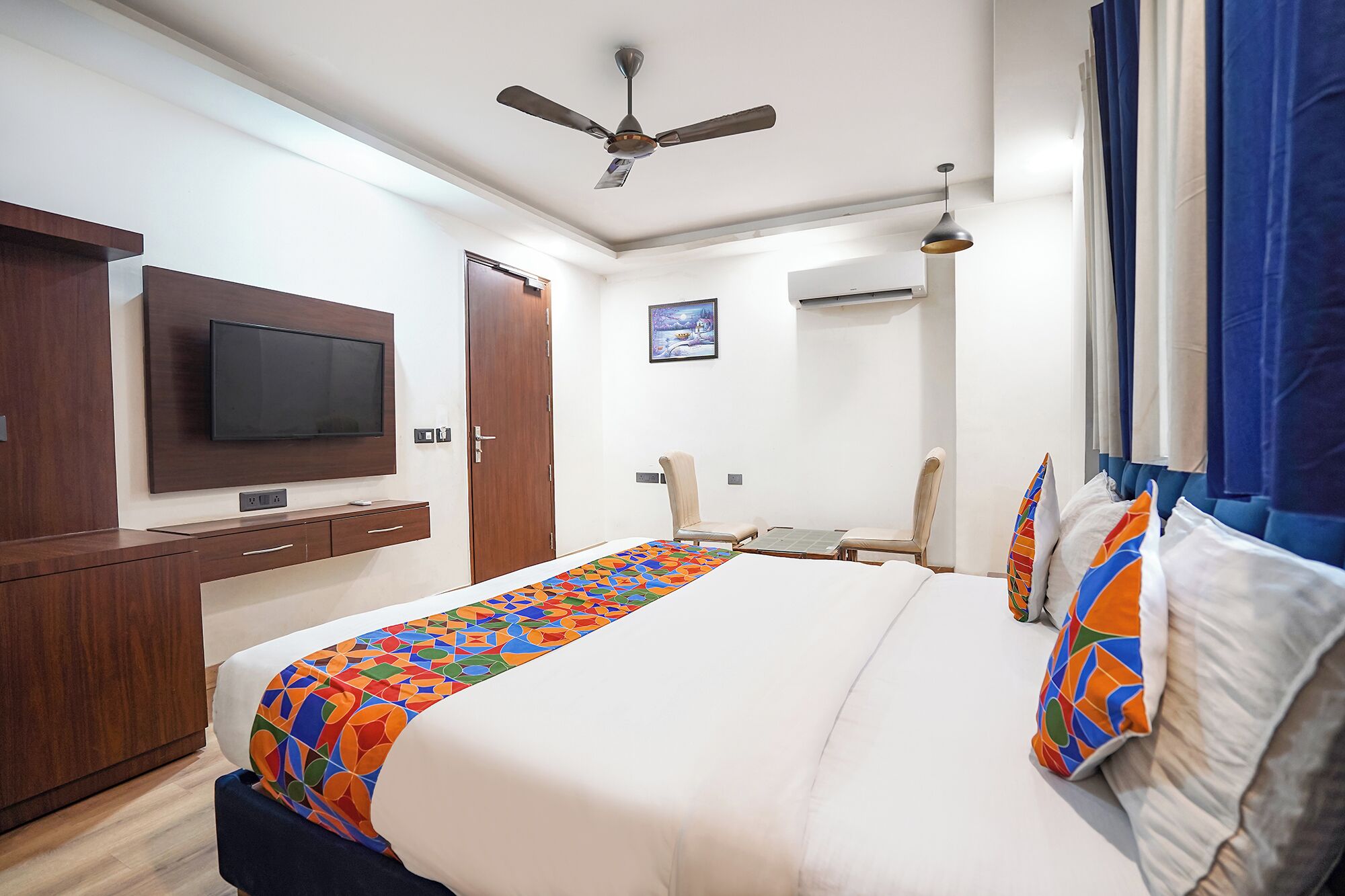 A hotel room with a bed, a TV mounted on the wall, a ceiling fan, an air conditioning unit, and a window with curtains.