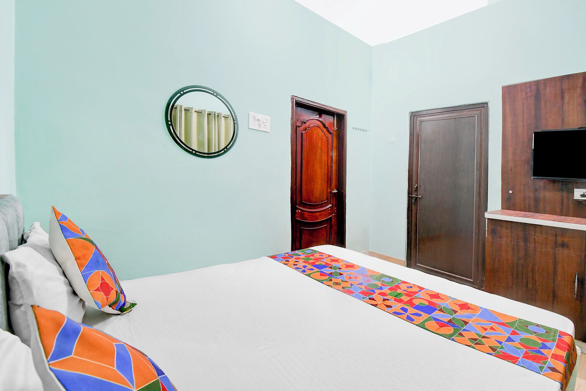 Un dormitorio con cama, espejo, puerta y televisor instalado en un mueble de madera.