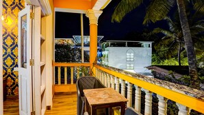 A balcony with a wooden table and chairs, a yellow railing, and a view of a building and palm trees at night. - Fabhotel Romeo’S (Calangute)