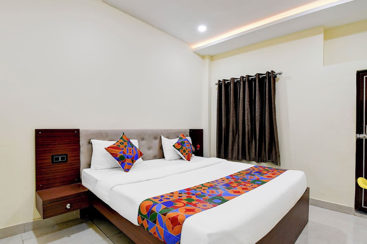 A neatly made bed with a colorful geometric-patterned bedspread and two pillows, a wooden nightstand with a drawer, and a window with dark curtains.