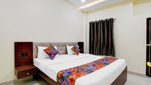A neatly made bed with a colorful geometric-patterned bedspread and two pillows, a wooden nightstand with a drawer, and a window with dark curtains. - Fabhotel Padmakar Kashi (Varanasi)