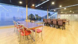 A spacious indoor area with wooden flooring, multiple tables and chairs, and large windows offering a view of the outdoors. - Fabhotel Burans Residency (Dhanaulti)