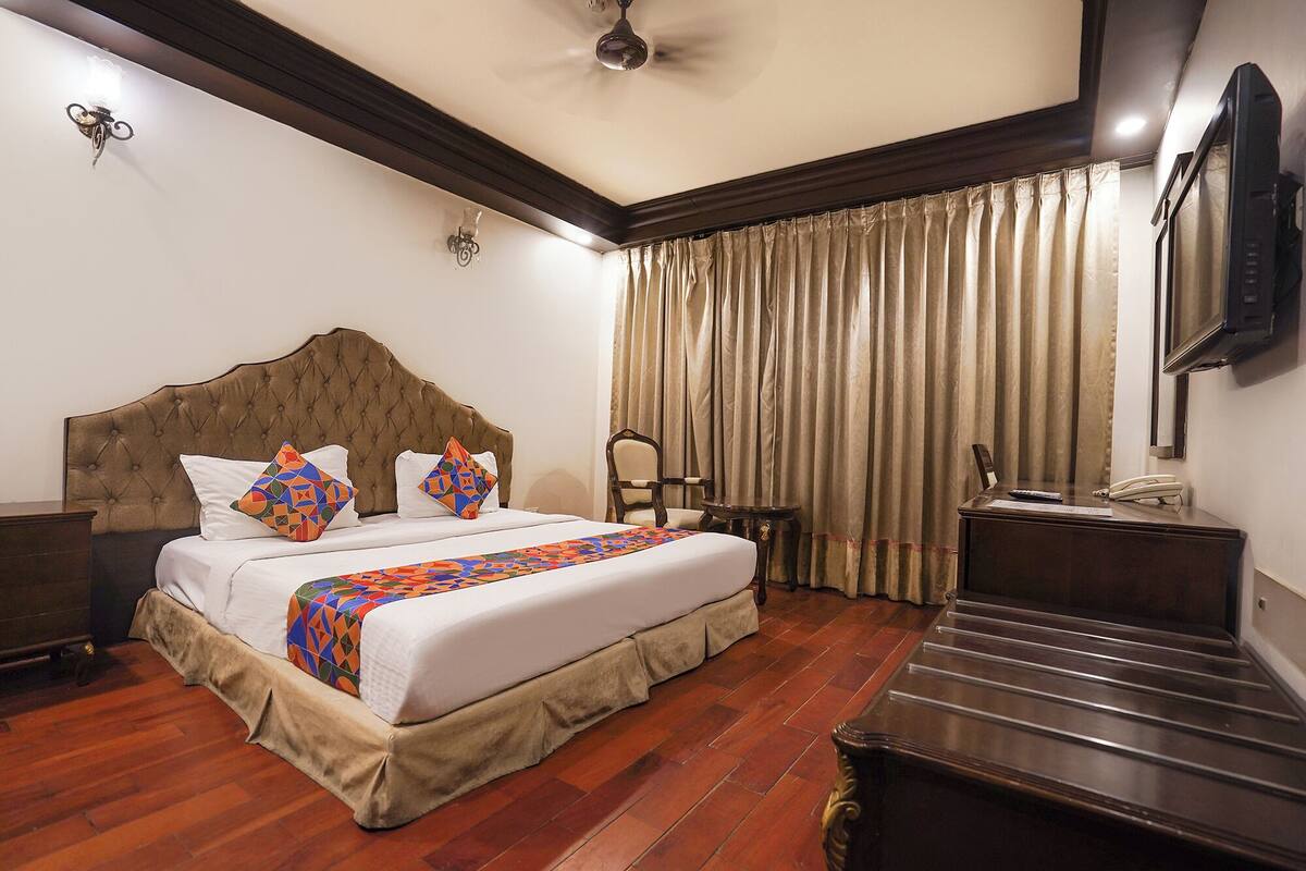 A hotel room with a tufted headboard bed, a wooden floor, a desk with a telephone, and a television on the wall.