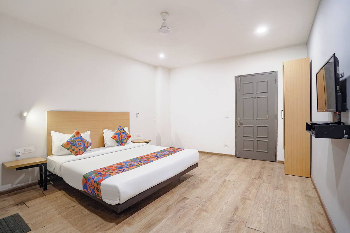 Quarto de hotel com cabeceira de madeira, cama com almofadas coloridas, mesa de cabeceira, televisão de tela plana na parede e porta de madeira.