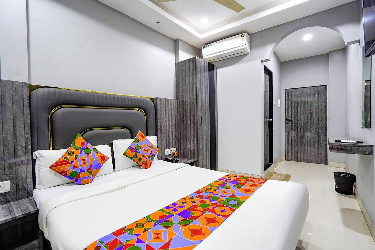 A modern hotel room with a large bed, a headboard, a bedside table, a nightstand, an air conditioning unit, a bathroom with a mirror, and a door.